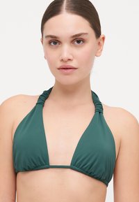 Tumši zaļš halter stila bikini topiņš ar savilkta auduma detaļu pie kakla, trīsstūra formas krūšu kausiem un elastīgu joslu zem krūtīm. Gluda, matēta tekstūra.
