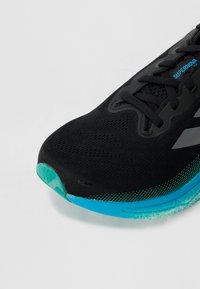 Scarpa sportiva nera con tomaia testurizzata, caratterizzata da una suola turchese e da sottili accenti a gradiente. Include lacci e dettagli del logo.