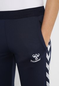 Marineblaue Sporthose aus einem glatten, dehnbaren Stoff. Verfügt über ein weißes Logo und Chevron-Muster entlang der seitlichen Nähte. Elastischer Bund.