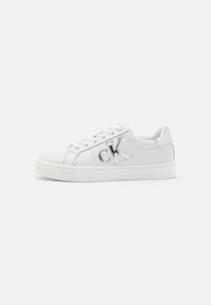 Zapatilla blanca de caña baja con logo plateado "ck" en el lateral, cordones blancos y suela plana, mostrada sobre un fondo claro y liso.