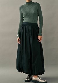 Pull à col roulé côtelé vert foncé associé à une jupe noire volumineuse et des chaussures argentées métalliques. Texture douce et forme fluide.