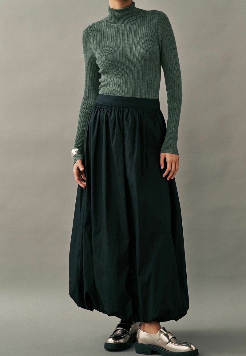 Pull à col roulé côtelé vert foncé associé à une jupe noire volumineuse et des chaussures argentées métalliques. Texture douce et forme fluide.