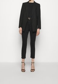 Svart skräddarsydd kavaj med dubbelknäppt design, bärs över en figurnära svart topp, ihop med smala svarta byxor och svarta sandalettklackar.