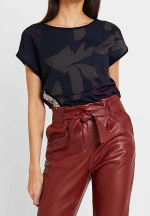 Frau trägt ein schwarz gemustertes Kurzarm-Top, das in hoch taillierte, bordeauxrote Lederhosen mit gebundenem Gürtel gesteckt ist.