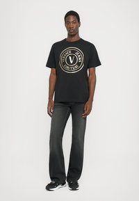 Tricou negru cu un mare logo circular aurit, care indică "VERSACE JEANS COUTURE" și un "V" în centru, asortat cu blugi gri închis și adidași negri.