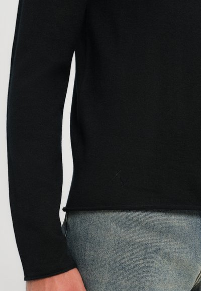 Black knit sweater with long sleeves, ribbed hem, and subtle branding detail. Worn over light blue denim, showcasing texture contrast.