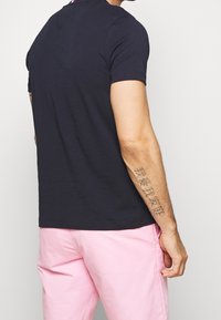 T-shirt blu navy a maniche corte con colletto rotondo, realizzato in cotone, mostrato da dietro, abbinato a pantaloni rosa chiaro, che mette in mostra un tatuaggio sul braccio.