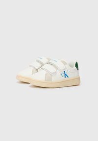 Vita sneakers med blå "Calvin Klein"-logga, kardborreband, beige detaljer och grön häl, på en ljus gummisula.