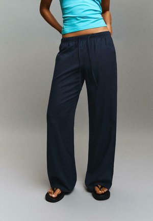 Person iført løse marineblå bukser, teal ærmeløs top og sorte klip-klappere, stående mod en ensfarvet grå baggrund.