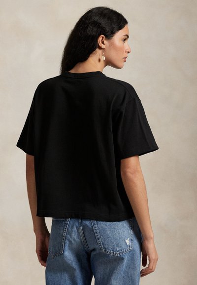 Naine pikkade tumedate juustega, kandes lahtist musta lühikese varrukaga särki ja siniseid teksapükse, külje pealt neutraalse tausta ees.