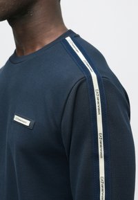 Muž, ktorý má na sebe námornícky modrú košeľu s biela a námorníckou modrou lemovkou na ramene a nášivkou na hrudi s nápisom "EA7 Emporio Armani."