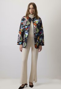 Giubbotto trapuntato con motivi floreali colorati su sfondo nero, dotato di colletto, bottoni frontali e due tasche. Indossato sopra pantaloni beige.