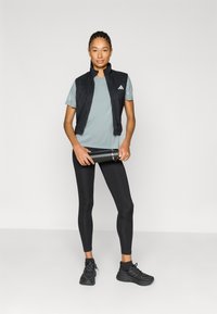 Czarna kamizelka bez rękawów, szary t-shirt, czarne legginsy i czarne trampki; trzyma czarną butelkę na wodę; gładka faktura, sportowy krój.