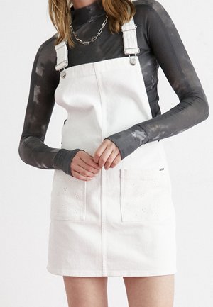 Femme portant une robe salopette en jean blanc sur un t-shirt à manches longues noir et gris tie-dye, mains jointes devant.