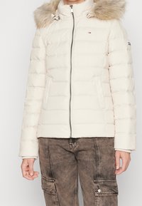 Pehmeä, vaniljainen toppatakki, jossa on korkea kaulus, vetoketjullinen suljenta ja irrotettava tekoturkisvuori. Takissa on sivutaskut ja hihassa yrityksen logo.