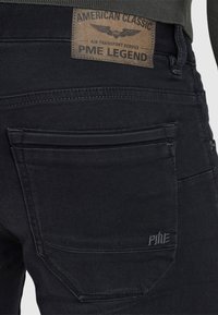 Vista posteriore di jeans in denim nero con tasca posteriore visibile e toppa in pelle marrone con l'etichetta "PME Legend American Classic."