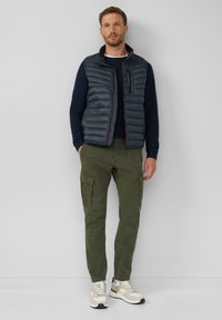 Herenoutfit met een donkergrijze puffervest, marineblauwe trui, olijfgroene cargobroek en beige sneakers met witte veters en zwarte accenten.