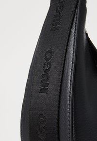 Borsa nera a spalla con corpo in pelle testurizzata e tracolla in tessuto impressa con "HUGO". Elementi di design concisi e moderni.