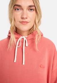 Coral hoodie with a drawstring, featuring a soft texture and a small logo on the chest. The material appears cozy and casual.