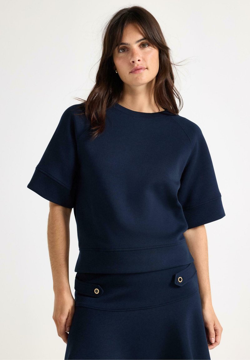 Navyblå sweatshirt i mjuk tyg, korta ärmar och vid halsringning. Har en figursydd midja och dekorativa detaljer på ärmsluten.