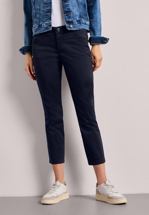 Kvinde iført korte marineblå bukser, hvide sneakers, hvid top og en denimjakke, der står op ad en ensfarvet lys væg.