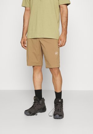Mann trägt beige Shorts und schwarze Wanderschuhe, steht vor einem schlichten weißen Hintergrund und trägt ein hellgrünes Hemd.