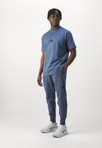 Niebieska bawełniana koszulka sportowa z krótkimi rękawami, z małym czarnym logo. W połączeniu z pasującymi niebieskimi spodniami dresowymi i białymi sneakersami.