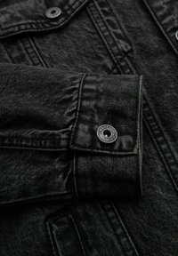 Dettaglio di una giacca in denim nera che mostra il tessuto testurizzato, le cuciture a punto e un bottone in metallo con testo inciso, che completa il design complessivo.