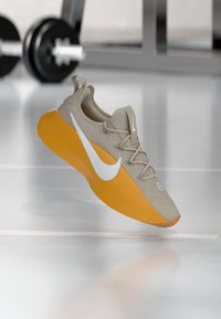 Chaussure de sport beige avec une semelle orange, des accents blancs, une tige texturée et des lacets. Présente un logo sur le côté et un col rembourré.