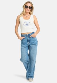 Hvid cropped tanktop med blå skrift, sat sammen med løstsiddende lyseblå jeans. Modellen bærer sorte solbriller og står med hænderne i lommerne.