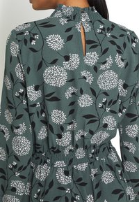 Robe fleurie verte avec un col smocké, une fermeture à boutons au dos et une taille élastique. Les motifs principaux sont des fleurs blanches et des feuilles noires.