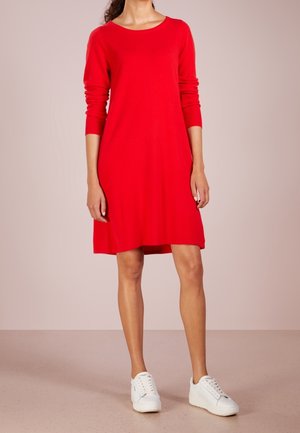 Vrouw draagt een rood langärmelig jurkje en witte plateau sneakers, staand tegen een lichtroze achtergrond.