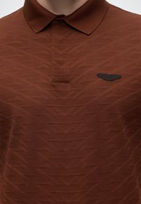 Gros plan d'une personne portant un polo marron texturé avec un emblème noir en forme d'aile sur la poitrine gauche.