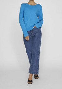Pull en maille bleu avec un col rond, associé à un jean en denim bleu taille haute. Sandales ouvertes foncées, coupe décontractée.