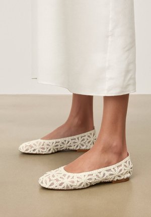 Fødder iført hvide blomsterhæklet ballet flats med en transparent mesh-base, kombineret med et hvidt midinederdel, stående på et glat gulv.