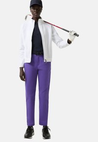 Bílá bunda na zip přes tmavě modrý top, v kombinaci s fialovými kalhotami a černými sportovními botami. Drží golfovou hůl s bílou rukavicí na jedné ruce.