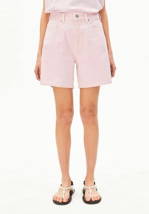 Persoon draagt lichtroze denim shorts met hoge taille, witte top en zwarte sandalen met witte gevlochten bandjes en staat tegen een effen witte achtergrond.