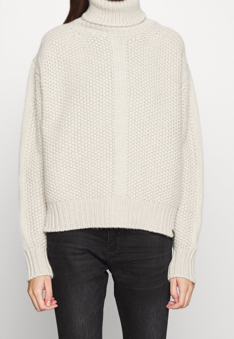 Gruby kremowy sweter z dzianiny z wysokim golfem, opuszczonymi ramionami, prążkowanymi mankietami i krawędzią. Posiada teksturowany wzór i luźny krój.