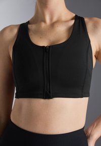 Svart sportsbh laget av elastisk materiale, med glidelås foran, brede stropper og en kort design. Glatt tekstur med sømdetaljer.