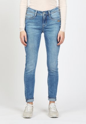 Vrouw draagt lichtblauwe opgerolde skinny jeans, wit shirt met lange mouwen en witte vetersneakers, staande rechtop.