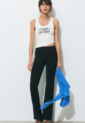 Femme portant un débardeur blanc avec le texte « Y a-t-il une raison que mon café ne soit pas là ? », pantalon noir, tenant un pull bleu contre un mur blanc.