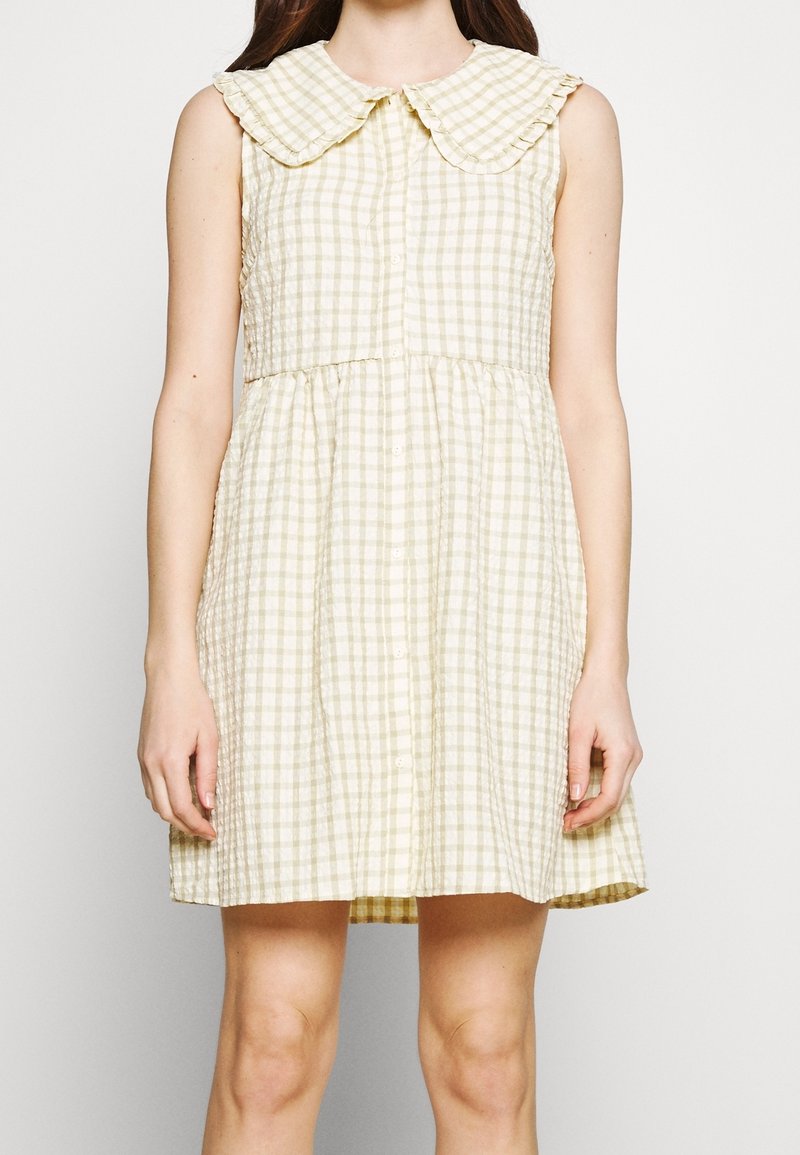 Robe sans manches beige et blanche à carreaux avec un grand col arrondi et des boutons devant, portée par une personne debout.