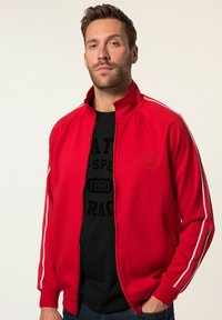 Giacca rossa con zip, strisce bianche ai lati, polsini a coste e logo in rilievo sul petto. Il tessuto sembra liscio e leggero.