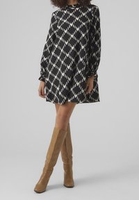 Kobieta ubrana w czarno-białą sukienkę w romby z długimi rękawami i brązowe skórzane botki do kolan, stojąca na tle gładkiej ściany.