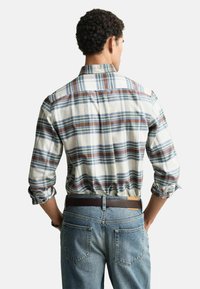 Camicia a quadri da uomo in tonalità bianche, blu e marroni, con colletto button-down e maniche arrotolate, abbinata a jeans blu in denim.