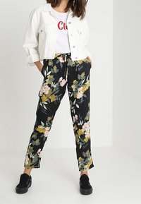 Pantalones negros con estampado floral en amarillo y rosa, con cintura con cordón, combinados con una chaqueta de mezclilla blanca cortada y zapatillas negras.