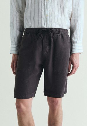 Hombre que lleva pantalones cortos oscuros con cordón y camisa de manga larga de color claro con finas rayas verticales, de pie frente a un fondo liso.