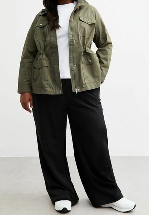 Personne portant une veste vert olive, une chemise blanche, un pantalon noir à jambes larges et des baskets blanches, debout devant un fond uni.