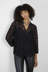 Schwarze Spitzenbluse mit langen Ärmeln, v-Ausschnitt und floralem Muster. Kombiniert mit glänzenden, schwarzen, figurbetonten Hosen.