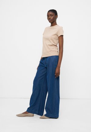 Femme portant un haut beige à manches courtes, un jean bleu à jambes larges et des ballerines beige, debout devant un fond blanc uni.